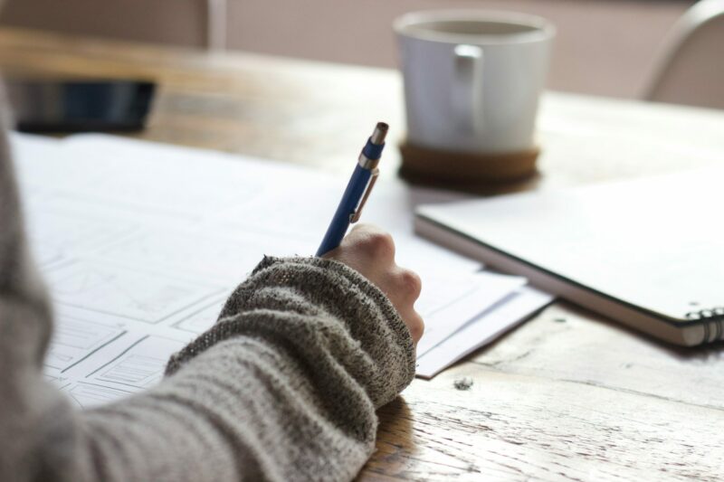 A hand draws on sheets of paper laying on a desk with a coffee cup just within reach