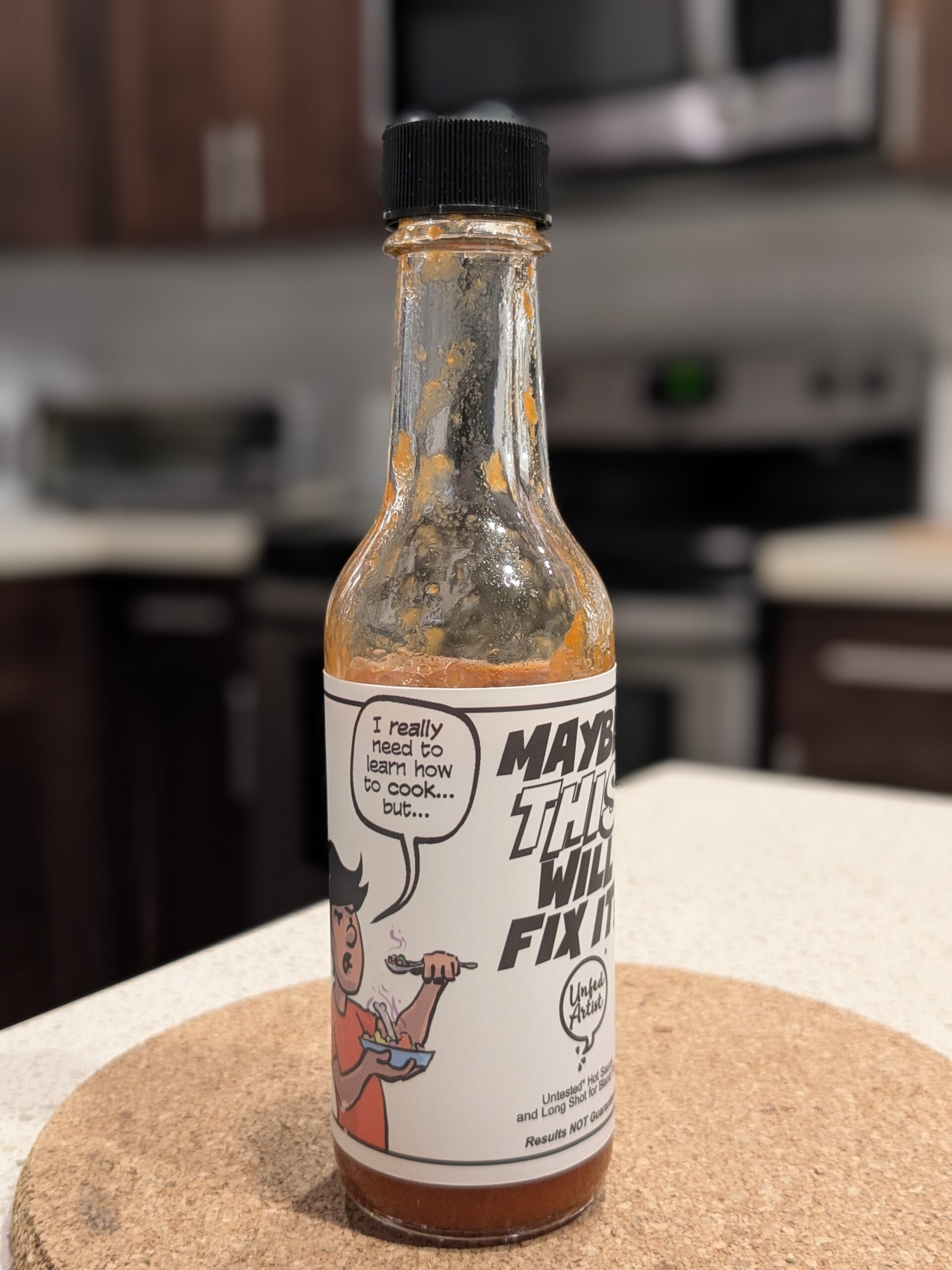 A half empty bottle of hot sauce with a label containing Ian's cartoon saying, "I really need to learn how to cook...but..." and the name of the sauce, "MAYBE THIS WILL FIX IT?"